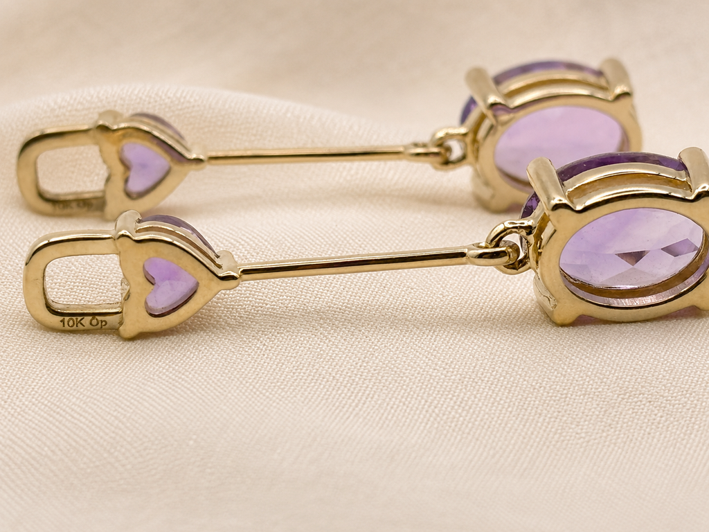 A pair of solid 10k yellow gold dangle earrings with oval purple amethyst stones by Ophelisse, displayed on a beige fabric background.