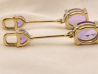 A pair of solid 10k yellow gold dangle earrings with oval purple amethyst stones by Ophelisse, displayed on a beige fabric background.