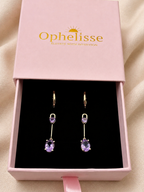 A pair of solid 10k yellow gold drop earrings with purple Amethyst gemstones displayed inside a pink Ophelisse gift box on a black insert, set against a soft beige fabric background.