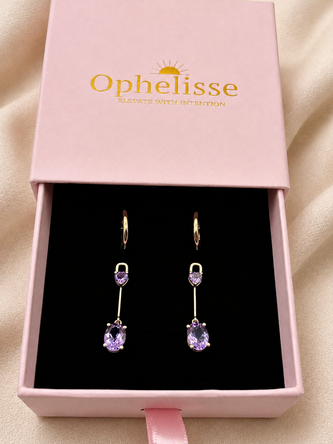 A pair of solid 10k yellow gold drop earrings with purple Amethyst gemstones displayed inside a pink Ophelisse gift box on a black insert, set against a soft beige fabric background.