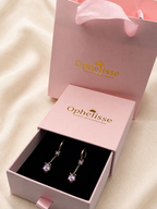 A pair of solid 10k yellow gold drop earrings with natural clear amethyst gemstones displayed in an open pink Ophelisse jewelry box, with the matching pink Ophelisse gift bag standing behind it on a soft cream fabric background.