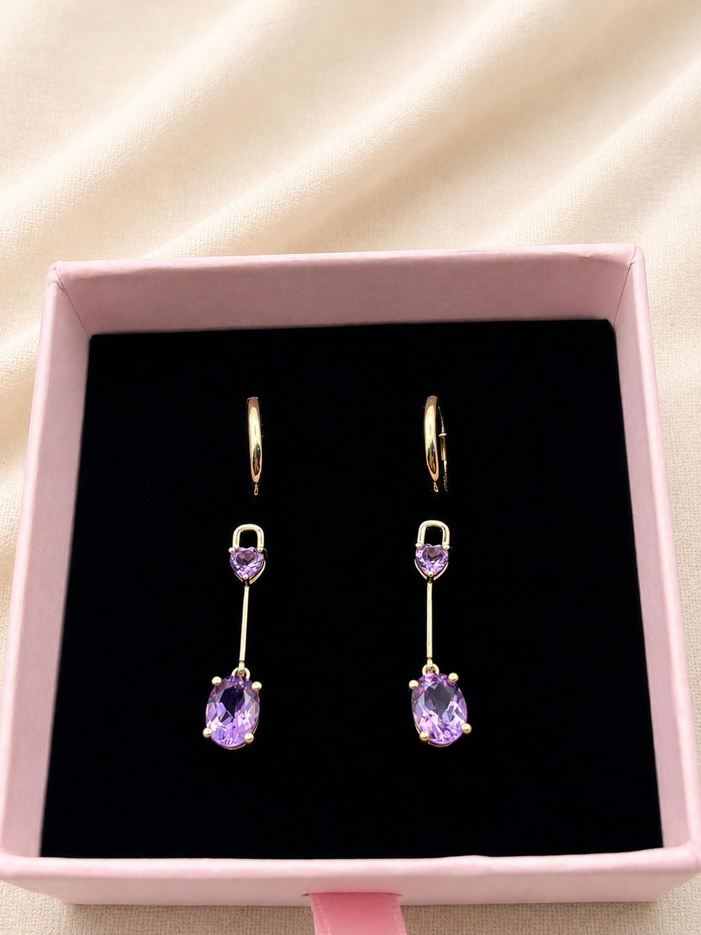 A pair of solid 10k yellow gold drop earrings with heart-shaped purple amethyst gemstones and oval purple drop stones by Ophelisse, displayed in a pink jewelry box with a black velvet lining.