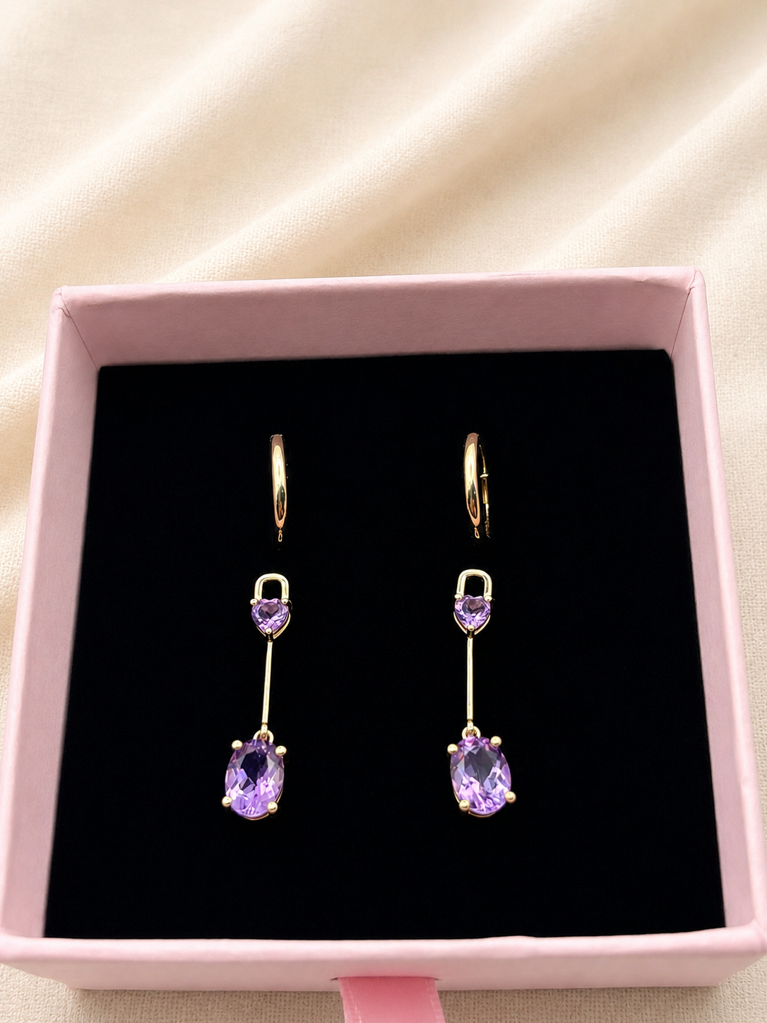 A pair of solid 10k yellow gold drop earrings with heart-shaped purple amethyst gemstones and oval purple drop stones by Ophelisse, displayed in a pink jewelry box with a black velvet lining.