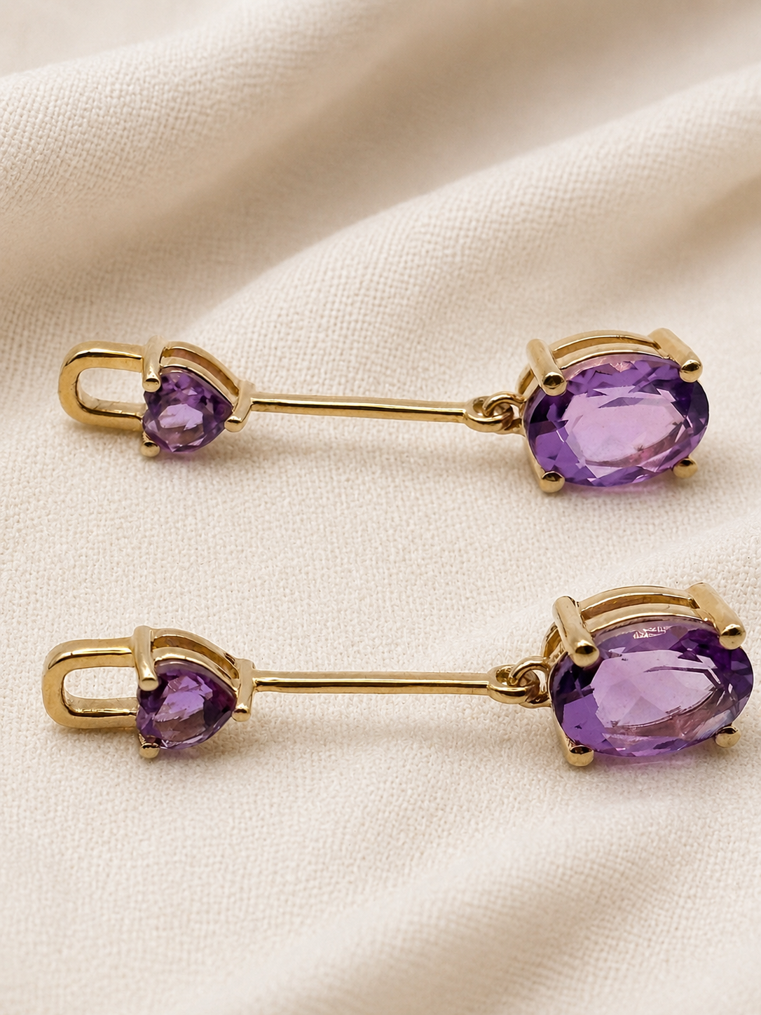A pair of solid 10k yellow gold drop earrings with small oval-shaped purple natural amethyst gemstones and larger oval purple natural amethyst gemstones by Ophelisse, displayed on a textured cream fabric background.