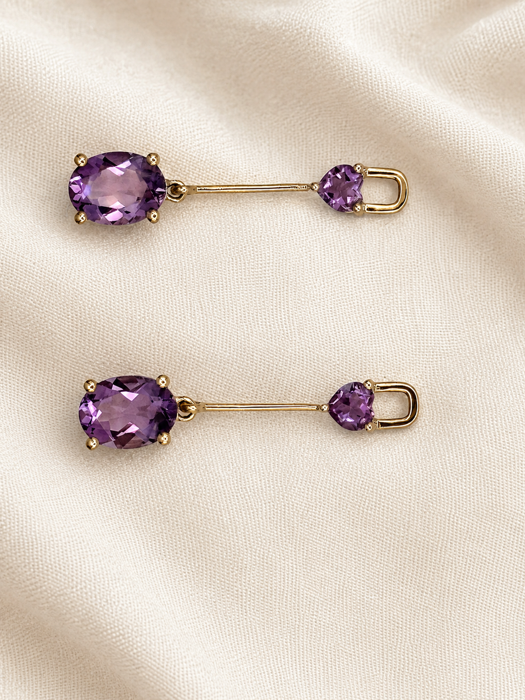 A pair of solid 10k yellow gold oval-style earrings with oval purple natural amethyst gemstones on one end and small oval-shaped purple natural amethyst gemstones on the other by Ophelisse, displayed on a cream-colored fabric background.