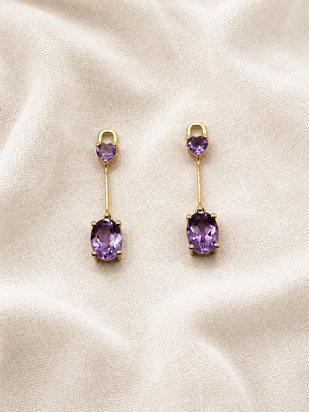 A pair of solid 10k yellow gold drop earrings with oval purple natural amethyst gemstones by Ophelisse, displayed on a textured beige fabric background.