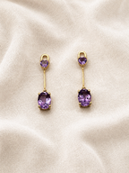 A pair of solid 10k yellow gold drop earrings with oval purple natural amethyst gemstones by Ophelisse, displayed on a textured beige fabric background.
