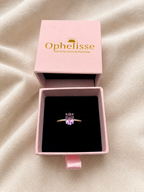 A solid 10k yellow gold ring with a purple oval-shaped amethyst gemstone displayed in a pink jewelry box with the brand name 'Ophelisse' on the lid, set against a beige fabric background.