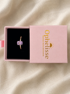A solid 10k yellow gold ring with a natural oval purple amethyst gemstone displayed in an open pink jewelry box with the brand name 'Ophelisse' printed on the lid, set against a draped cream fabric background.