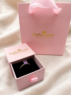 A solid 10k yellow gold ring with a natural purple amethyst gemstone displayed in an open pink Ophelisse jewelry box, with a matching pink Ophelisse shopping bag behind it on a soft beige fabric background.