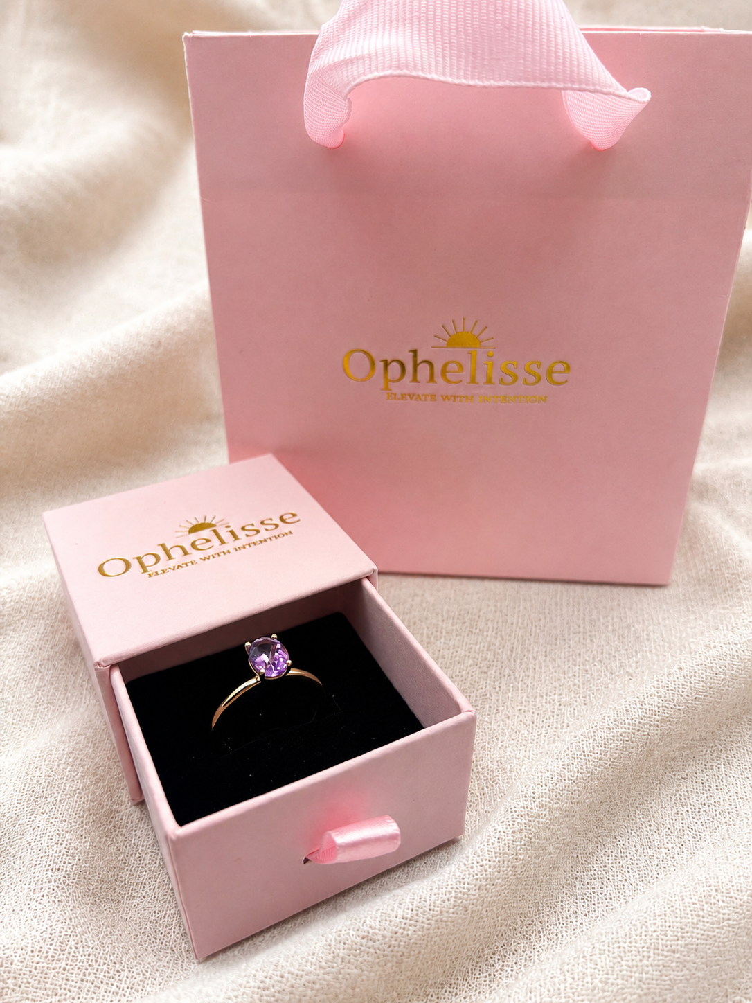 A solid 10k yellow gold ring with a natural purple amethyst gemstone displayed in an open pink Ophelisse jewelry box, with a matching pink Ophelisse shopping bag behind it on a soft beige fabric background.