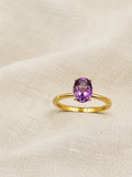 A solid 10k yellow gold ring with an oval natural purple amethyst gemstone by Ophelisse, resting on a textured beige fabric background.
