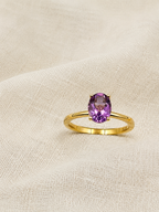 A solid 10k yellow gold ring with an oval natural purple amethyst gemstone by Ophelisse, resting on a textured beige fabric background.