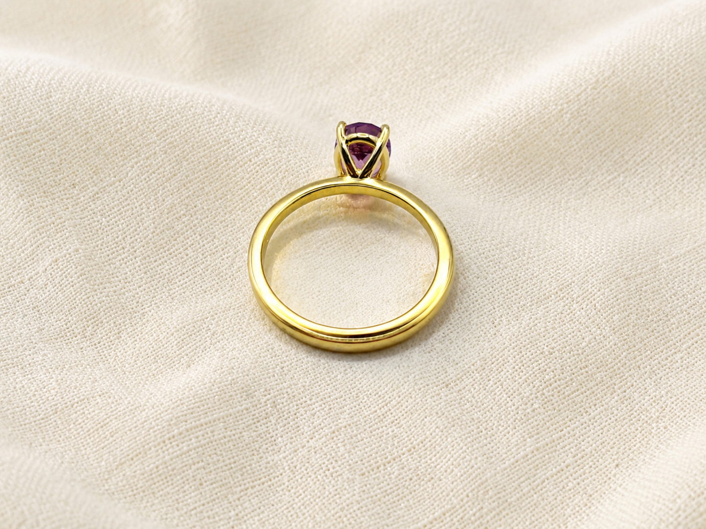 A solid 10k yellow gold ring with a small oval natural purple amethyst gemstone in a prong setting by Ophelisse, displayed on a textured beige fabric background.