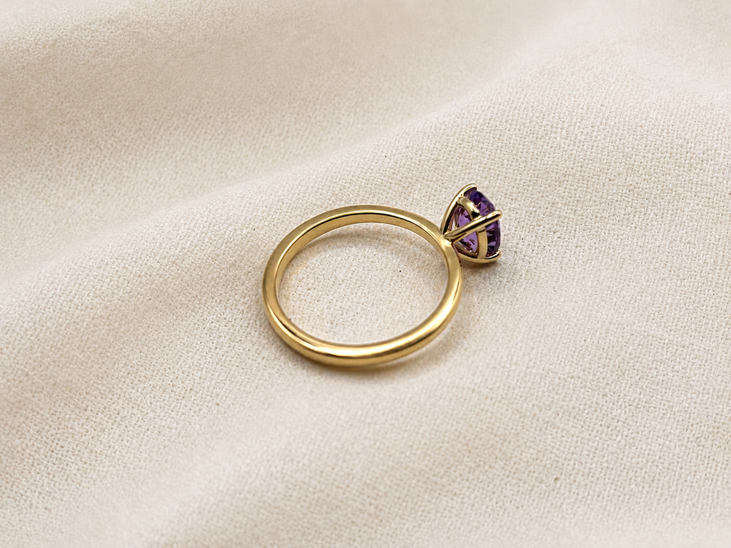 A solid 10k yellow gold ring with a oval natural purple amethyst gemstone in a prong setting by Ophelisse, resting on a textured beige fabric background.