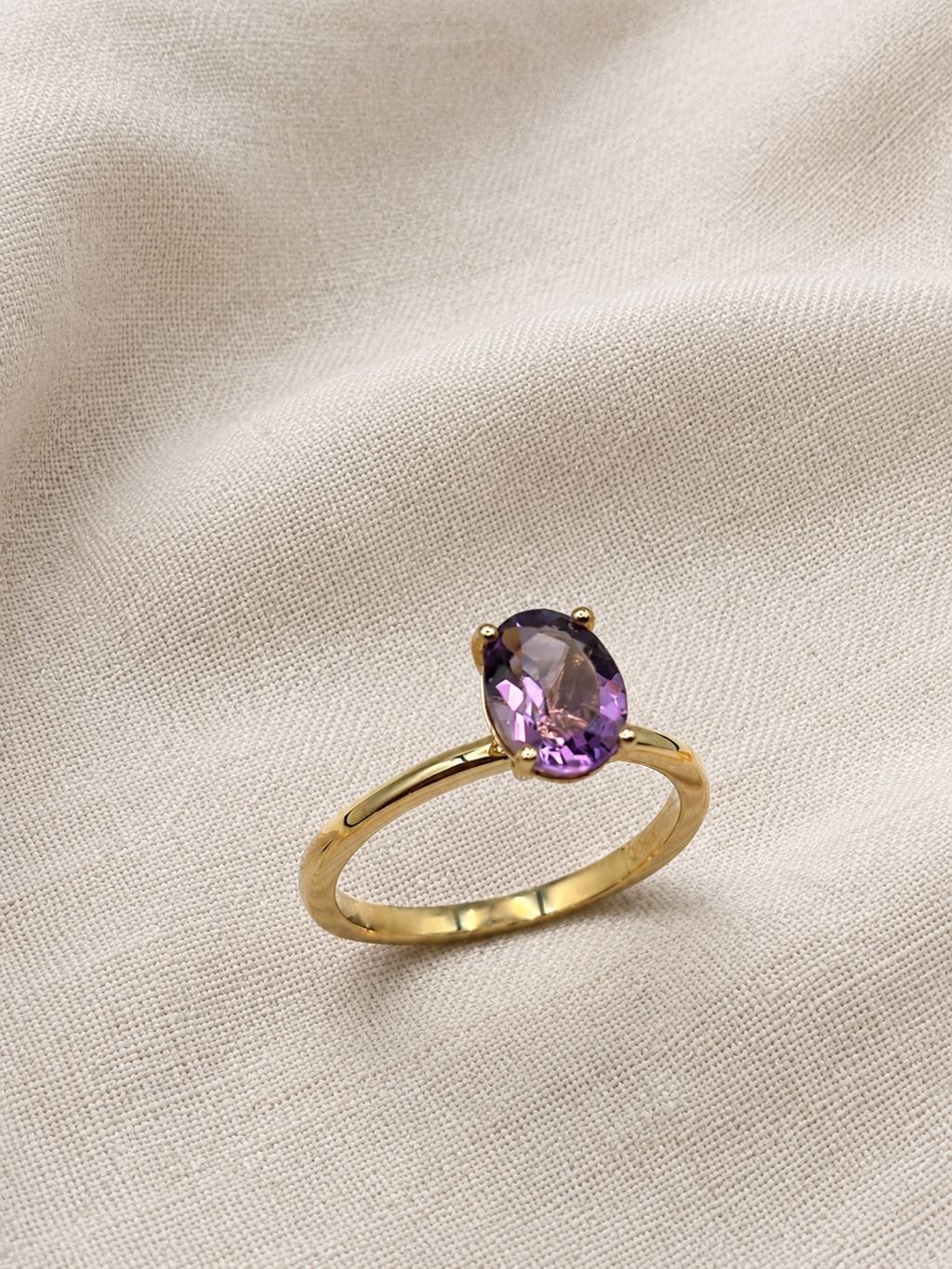 A solid 10k yellow gold ring with a oval-shaped natural purple amethyst gemstone by Ophelisse, resting on a textured beige fabric background.