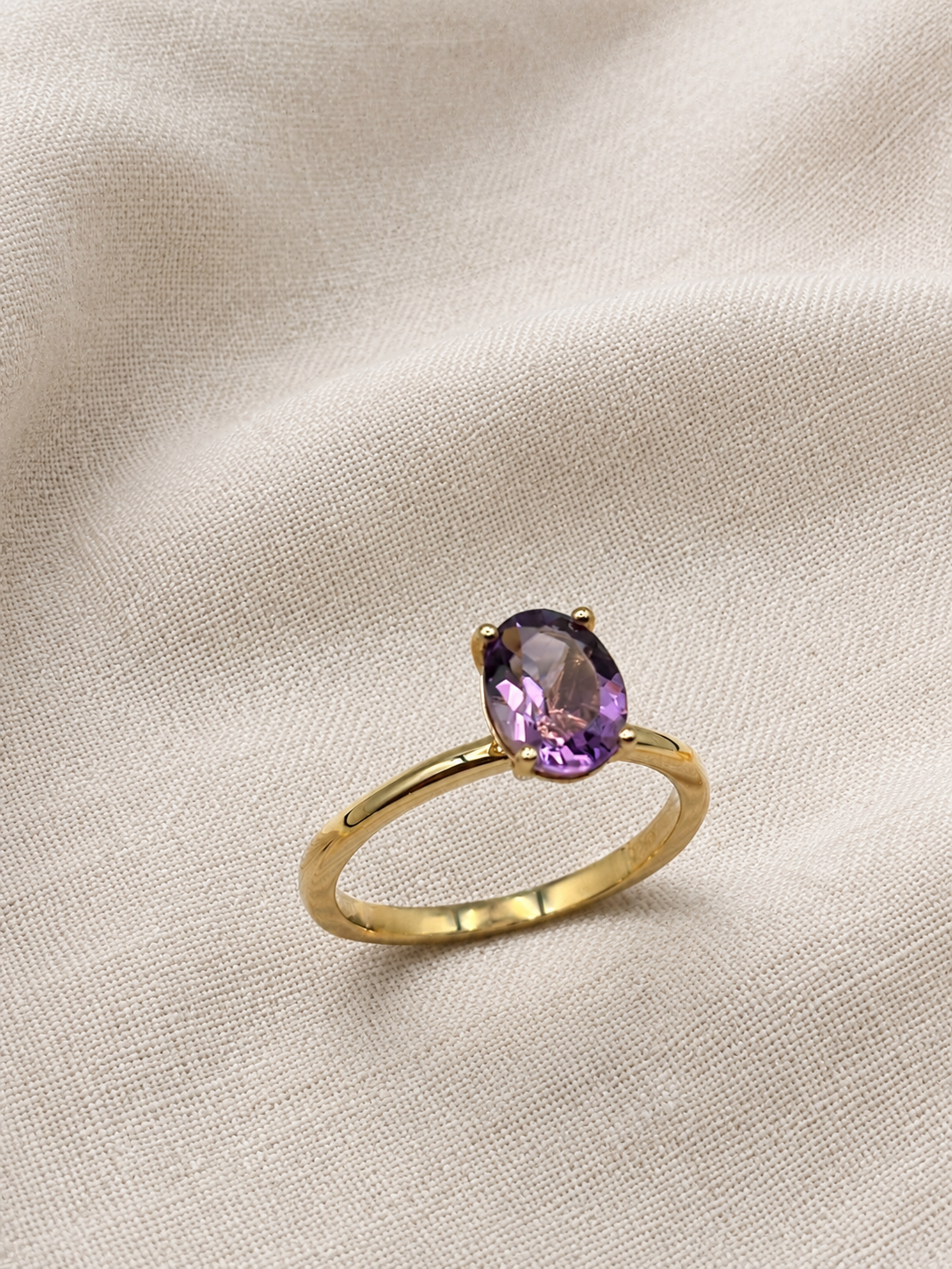 A solid 10k yellow gold ring with a oval-shaped natural purple amethyst gemstone by Ophelisse, resting on a textured beige fabric background.