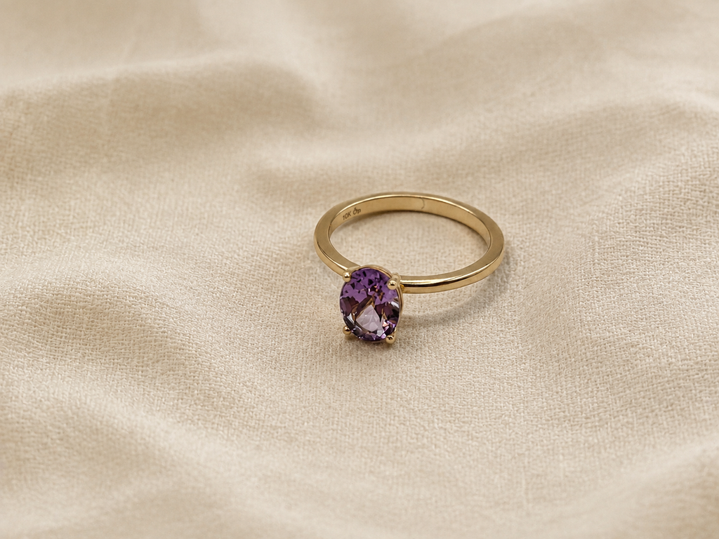 A solid 10k yellow gold ring with a oval natural purple amethyst gemstone by Ophelisse, resting on a textured beige fabric background.