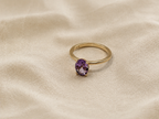 A solid 10k yellow gold ring with a oval natural purple amethyst gemstone by Ophelisse, resting on a textured beige fabric background.