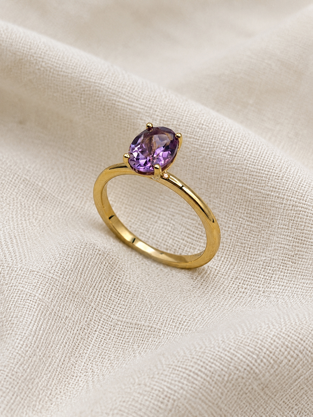 A solid 10k yellow gold ring with a oval-cut natural purple amethyst gemstone by Ophelisse, resting on a textured beige fabric background.