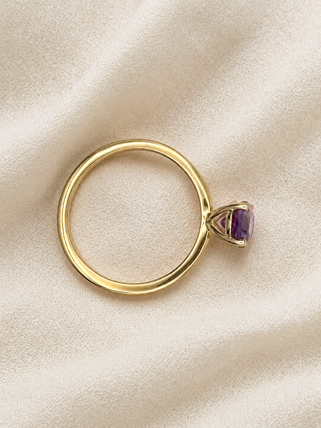 A solid 10k yellow gold ring with a small oval natural purple Amethyst gemstone by Ophelisse, displayed on a textured beige fabric background.