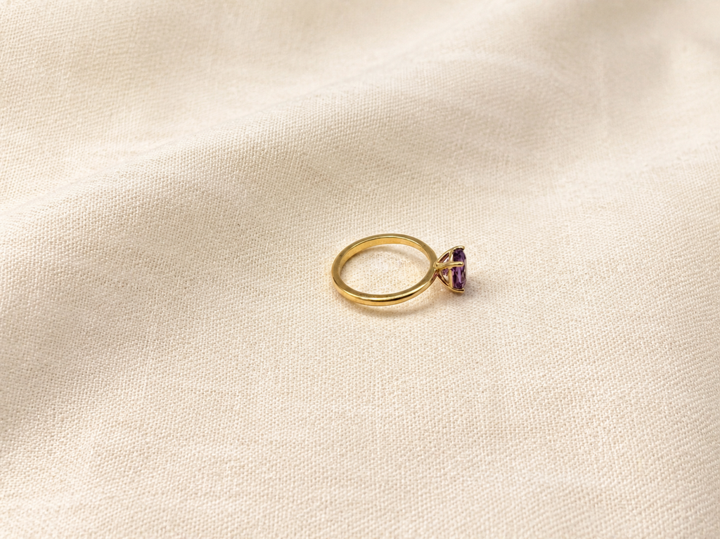 A solid 10k yellow gold ring with a oval natural purple amethyst gemstone by Ophelisse, resting on a textured beige fabric background.