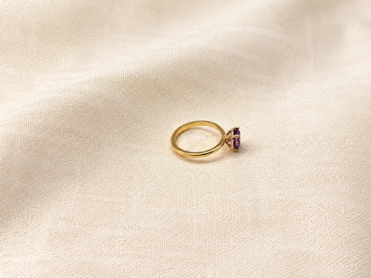 A solid 10k yellow gold ring with a oval natural purple amethyst gemstone by Ophelisse, resting on a textured beige fabric background.