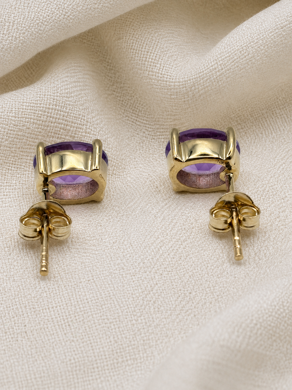 A pair of solid 10k yellow gold stud earrings with oval purple natural amethyst gemstones by Ophelisse, displayed on a textured cream fabric background.