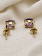A pair of solid 10k yellow gold stud earrings with oval purple natural amethyst gemstones by Ophelisse, displayed on a textured cream fabric background.