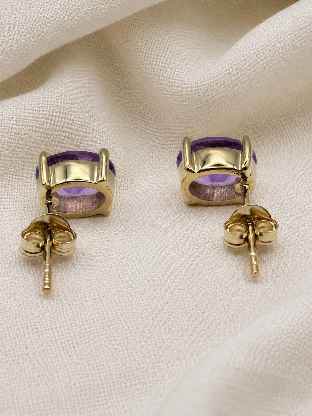 A pair of solid 10k yellow gold stud earrings with oval purple natural amethyst gemstones by Ophelisse, displayed on a textured cream fabric background.