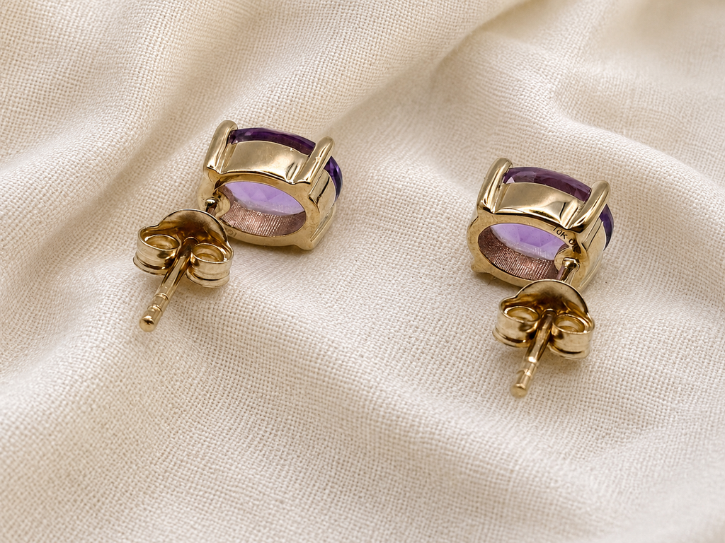 A pair of solid 10k yellow gold stud earrings with oval purple natural amethyst gemstones, displayed on a textured cream fabric background.