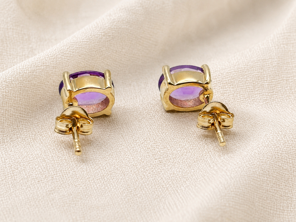 A pair of solid 10k yellow gold stud earrings with natural oval purple amethyst gemstones by Ophelisse, displayed on a textured beige fabric background.