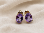 A pair of solid 10k yellow gold stud earrings with oval-cut purple natural amethyst gemstones, displayed on a textured beige fabric background.