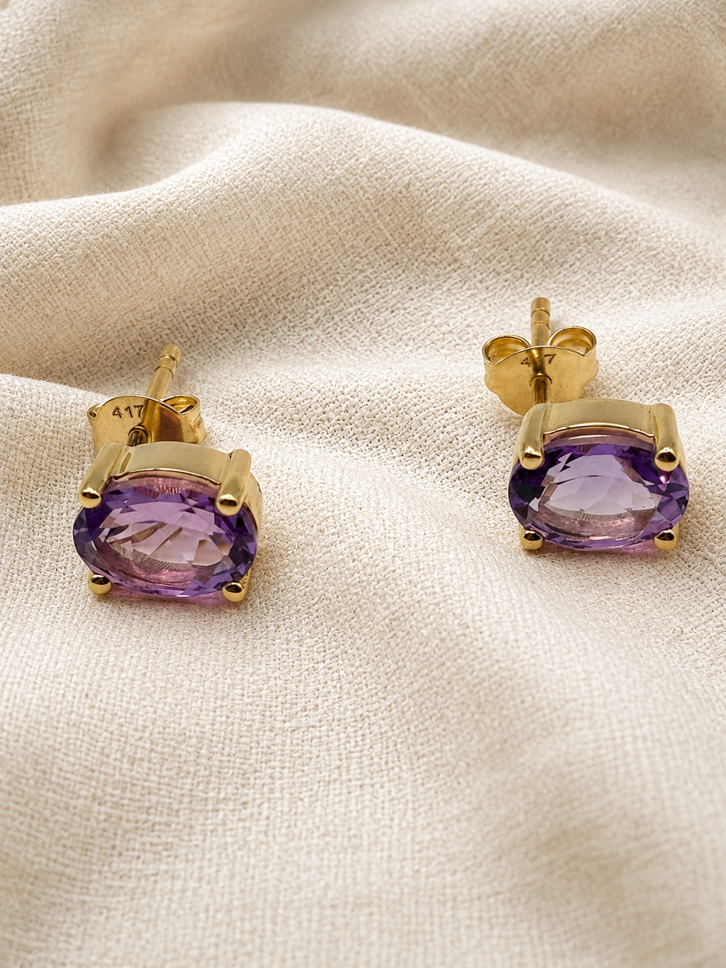 A pair of solid 10k yellow gold stud earrings with oval purple natural amethyst gemstones by Ophelisse, displayed on a textured beige fabric background.