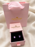 A pair of oval-cut purple natural amethyst gemstone stud earrings displayed in an open pink Ophelisse jewelry box, with the matching pink branded shopping bag standing behind it on draped cream fabric.