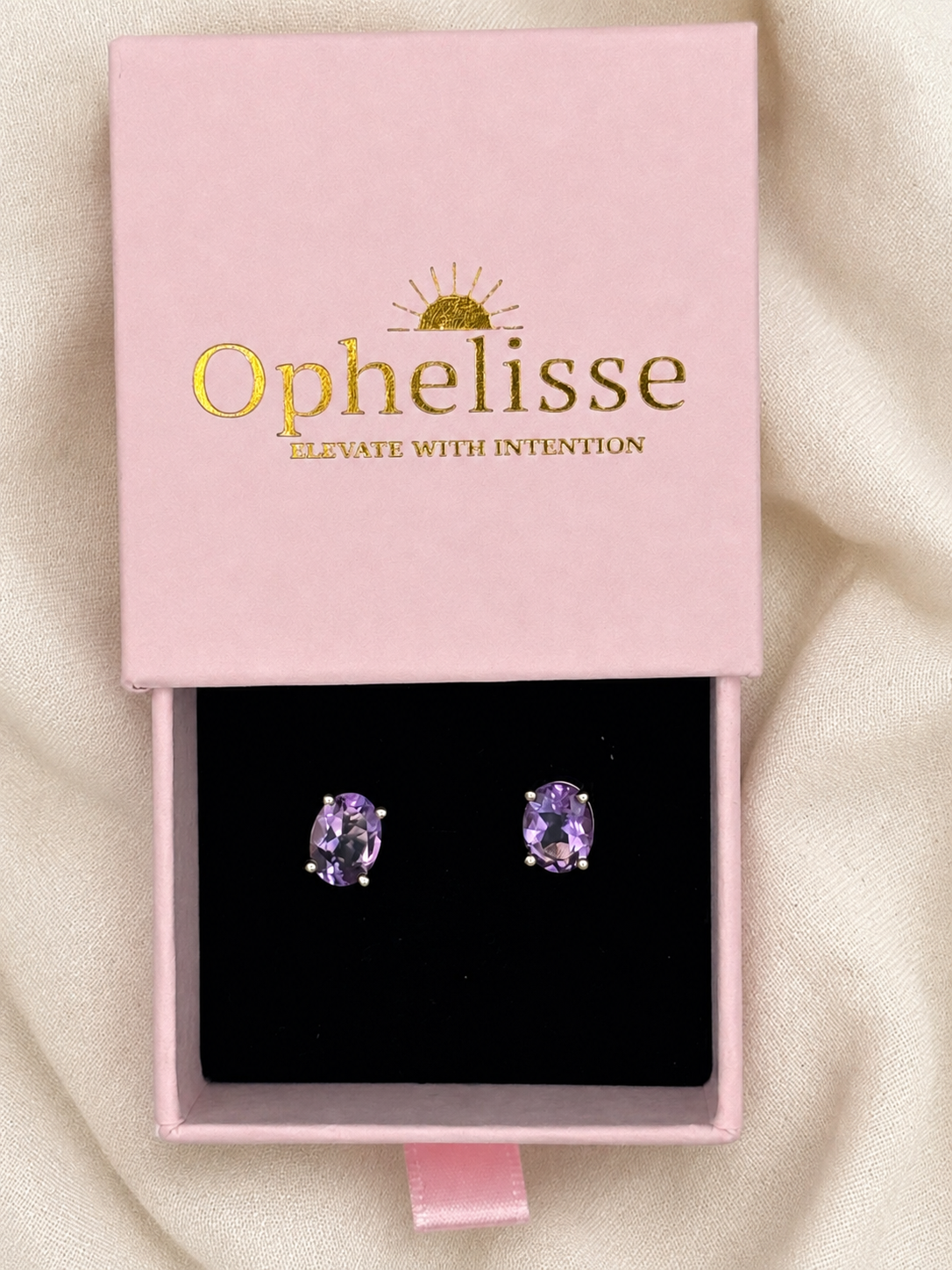 A pair of oval purple natural amethyst gemstone stud earrings displayed in a pink jewelry box with a black interior. The lid is printed with the brand name 'Ophelisse' in gold lettering with a small sun graphic above it.