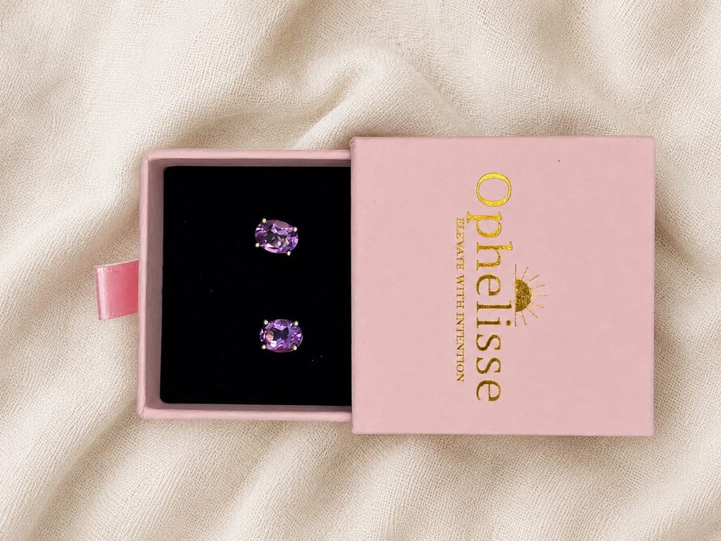 A pair of oval-cut natural purple amethyst gemstone stud earrings displayed in an open pink jewelry box with a branded pink lid, set on a cream fabric background.