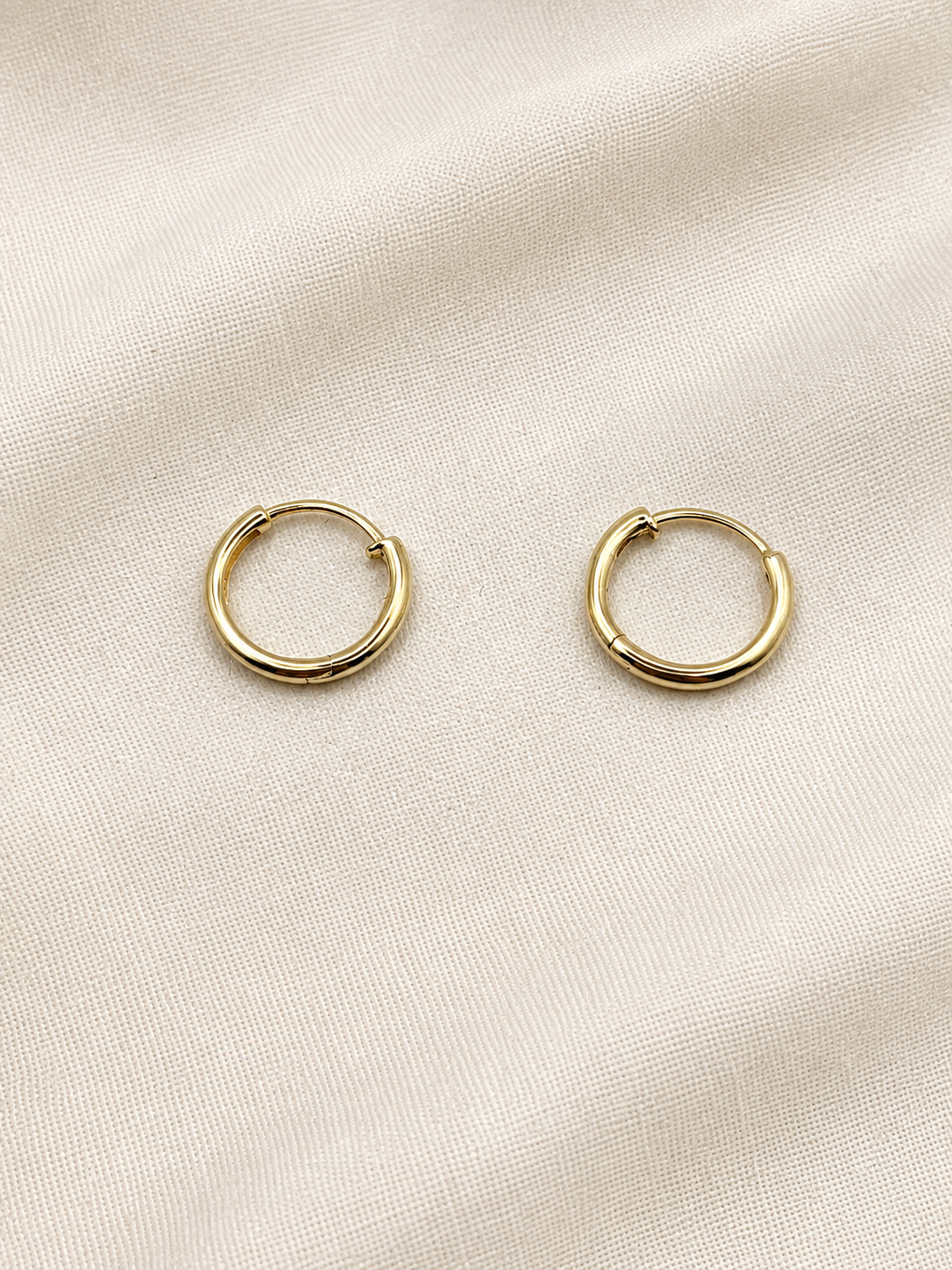 A pair of solid 10k yellow gold dangle earring hoops resting on a textured, light beige fabric background. Trademarked by Ophalisse.
