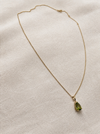 A delicate solid 10k yellow gold chain necklace with a small gold bezel holding a green natural peridot teardrop-shaped gemstone by Ophelisse, displayed on an elegant textured beige linen fabric background.