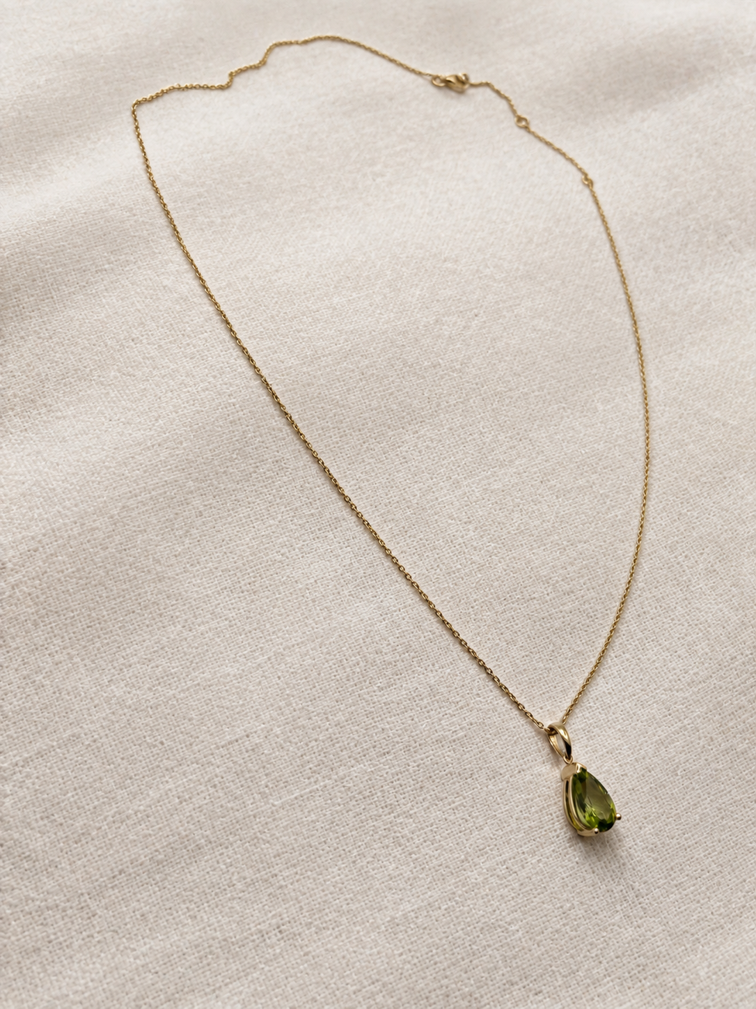 A delicate solid 10k yellow gold chain necklace with a small gold bezel holding a green natural peridot teardrop-shaped gemstone by Ophelisse, displayed on an elegant textured beige linen fabric background.