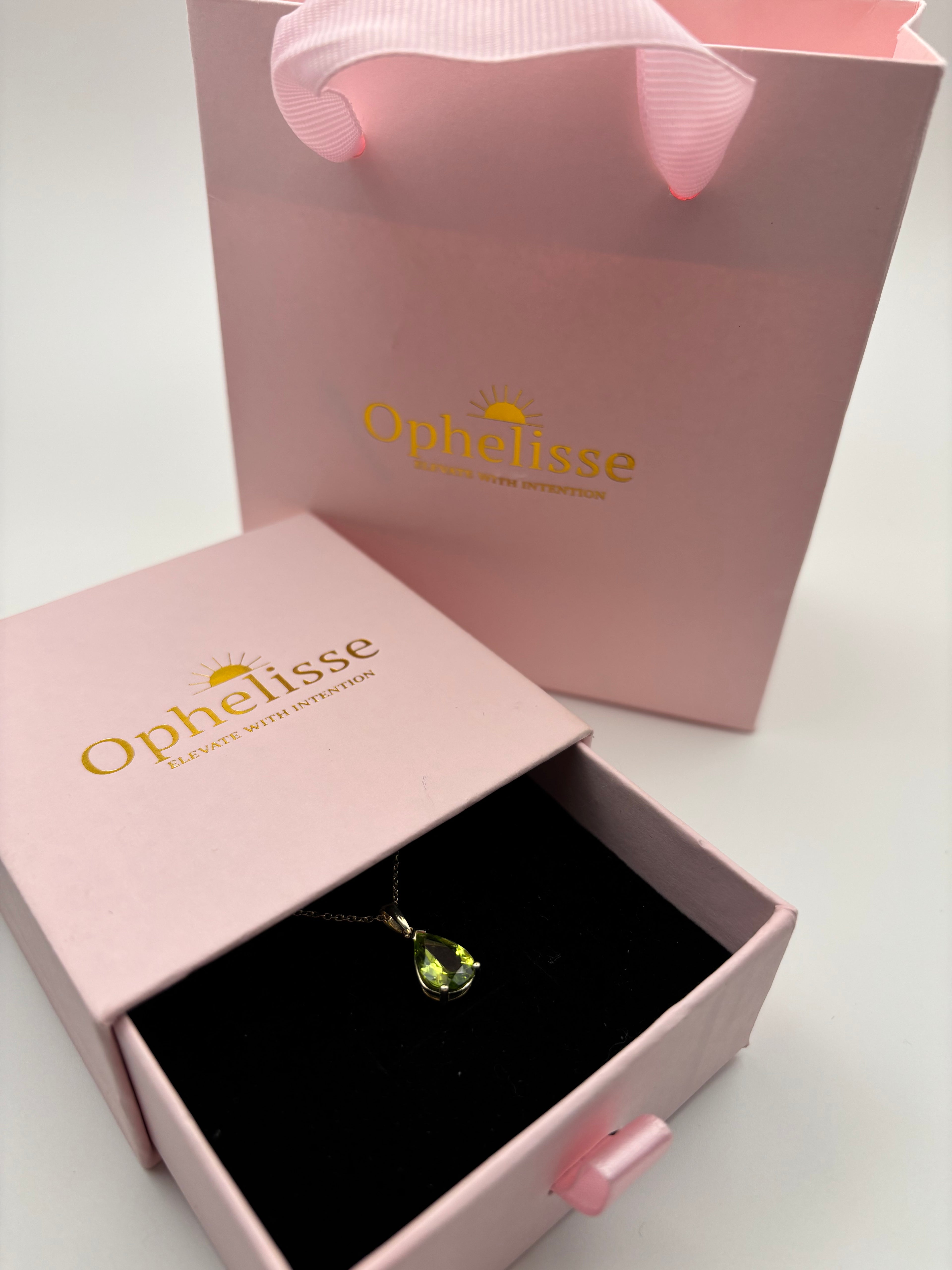 A 10k solid yellow gold pendant necklace with a green peridot teardrop natural gemstone displayed in an open pink luxurious Ophelisse jewelry box, with a pink beautiful branded shopping bag behind it on a white background.