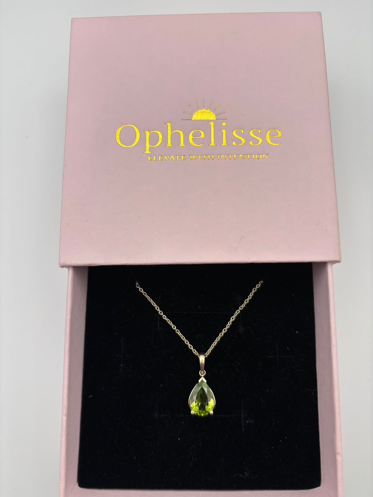 An elegant solid 10k yellow gold chain necklace with a teardrop-shaped green peridot natural gemstone pendant, presented inside an open pink jewelry box with the brand name 'Ophelisse' printed in gold on the lid.