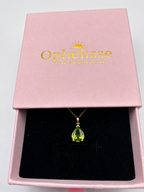A 10k solid yellow gold pendant necklace with a luxurious green peridot teardrop natural gemstone displayed inside an open elegant pink jewelry box with the brand name 'Ophelisse' printed in gold on the lid. Very good for momentous occasions and quiet time.