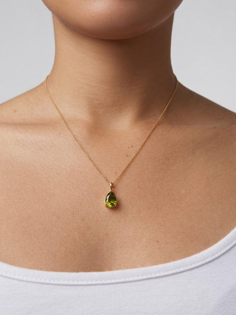 A close-up of a model's neck and upper chest wearing a delicate gold chain necklace with a small teardrop-shaped green gemstone pendant.