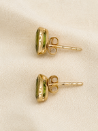 A pair of 10k yellow solid gold stud earrings with oval natural green peridot gemstones by Ophelisse, displayed on a textured beige fabric background.