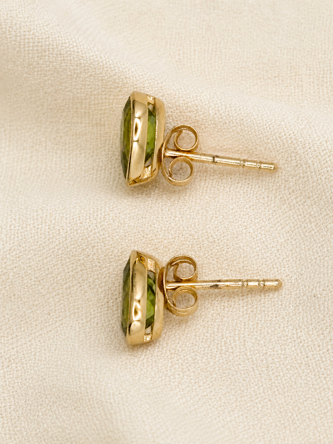 A pair of 10k yellow solid gold stud earrings with oval natural green peridot gemstones by Ophelisse, displayed on a textured beige fabric background.