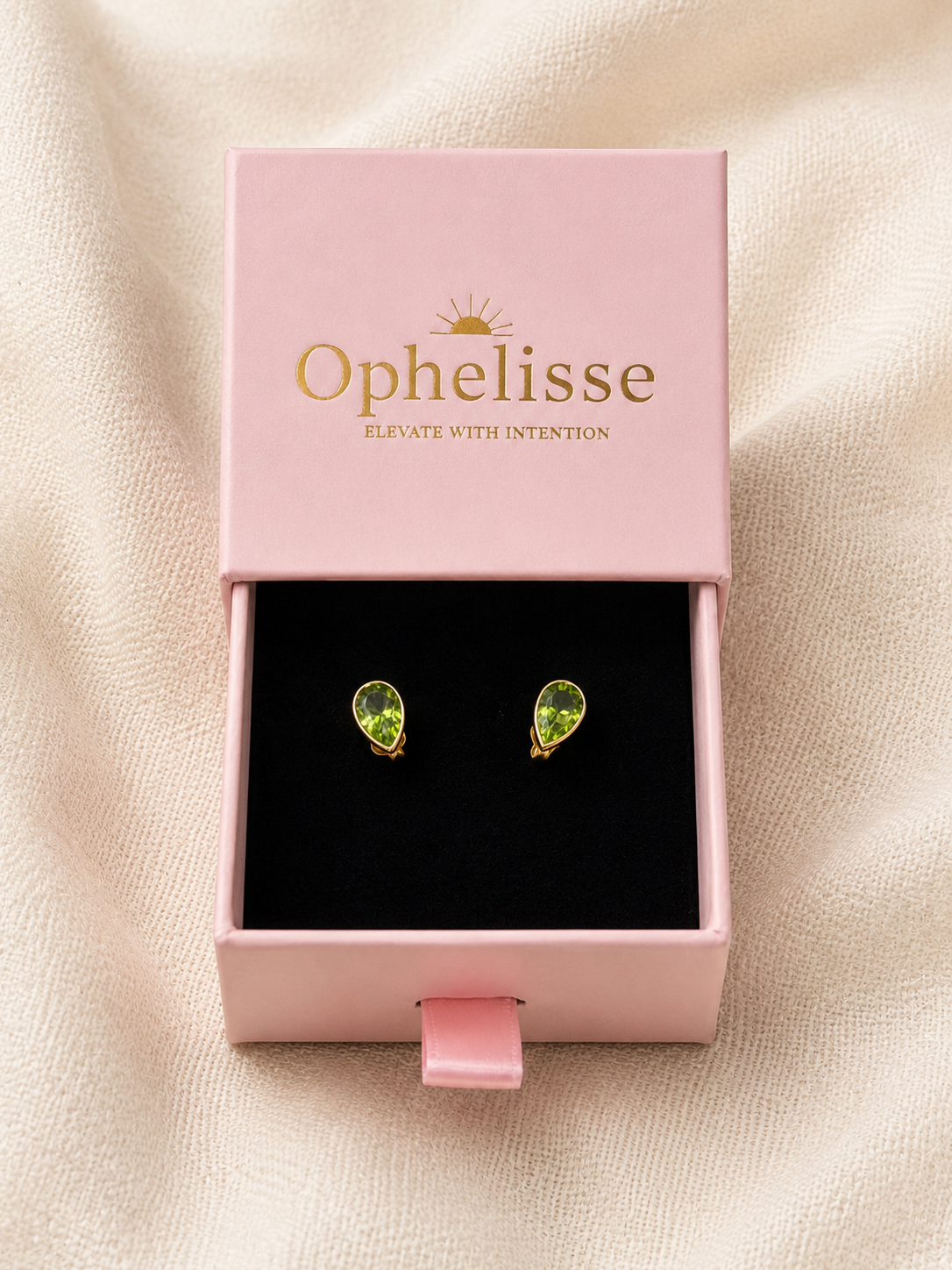 A pair of solid 10k yellow gold teardrop stud earrings with natural green Peridot gemstones displayed in an open pink Ophelisse jewelry box on a beige fabric background.