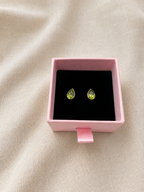 A pair of 10k solid yellow gold teardrop stud earrings with natural green peridot gemstones, presented in a small pink jewelry box on a beige fabric background.