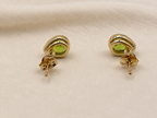 A pair of solid 10k yellow gold stud earrings with oval green peridot natural gemstones by Ophelisse, displayed on a textured beige fabric background with the back showing Ophelisse trademarks.