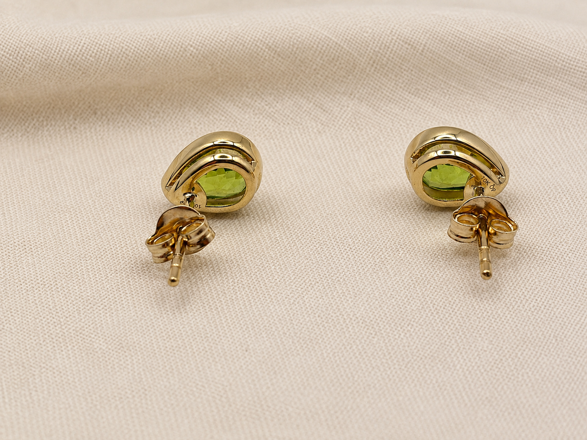 A pair of solid 10k yellow gold stud earrings with oval green peridot natural gemstones by Ophelisse, displayed on a textured beige fabric background with the back showing Ophelisse trademarks.